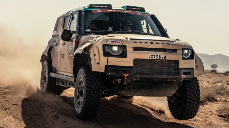 A Defender Dakar D7X-R mid-jump on a dusty desert stage, front bumper damaged from the terrain