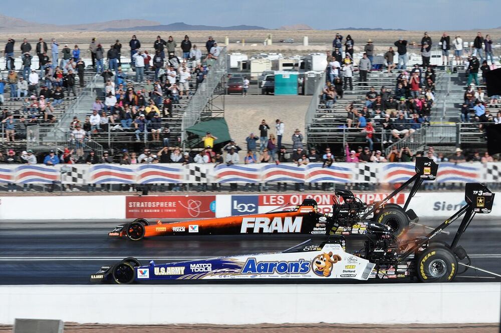 Two Top Fuel dragsters side by side at the start line in front of the grandstand